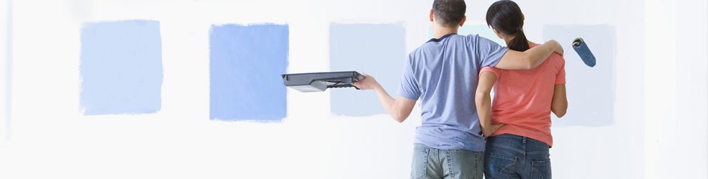 Couple holding paint roller and tray and examining blue paint samples on living room wall